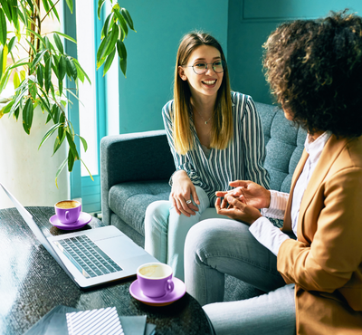 Two colleagues sitting on a couch discussing work with a laptop and two mugs on the coffee table.